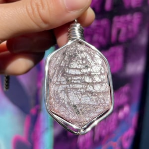 May include: A pendant featuring a faceted, pink gemstone encased in a silver wire frame. The gemstone has a textured surface with visible lines and dark inclusions. The wire wraps around the top, forming a loop for a chain. The background is blurred.