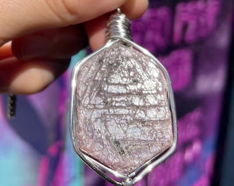 May include: A pendant featuring a faceted, pink gemstone encased in a silver wire frame. The gemstone has a textured surface with visible lines and dark inclusions. The wire wraps around the top, forming a loop for a chain. The background is blurred.
