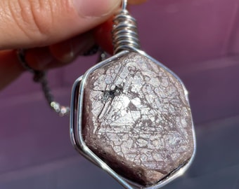 May include: A hexagonal, dusty rose-colored gemstone pendant encased in a silver-toned wire wrap. The stone features a textured, crystalline surface with visible geometric patterns and a metallic sheen, suspended from a silver-toned metal chain.