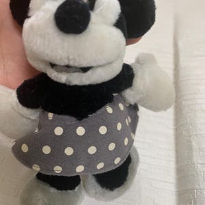 Op de afbeelding: Een Minnie Mouse pluche pop, met een zwart-wit kleurenschema. De pop heeft een grijze jurk met witte stippen, zwarte oren en een grijze hoed. Het gezicht is wit met zwarte ogen en een zwarte neus.