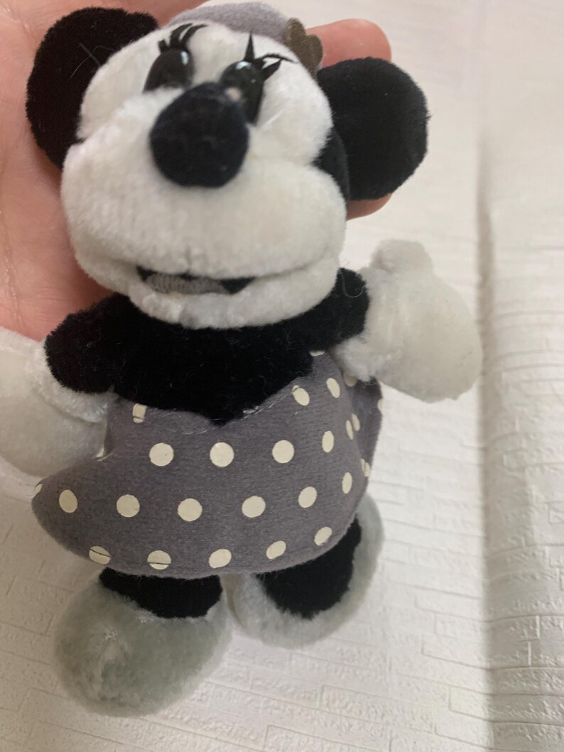 Op de afbeelding: Een Minnie Mouse pluche pop, met een zwart-wit kleurenschema. De pop heeft een grijze jurk met witte stippen, zwarte oren en een grijze hoed. Het gezicht is wit met zwarte ogen en een zwarte neus.
