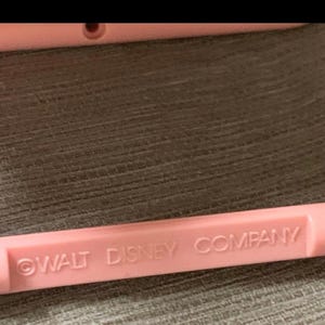 May include: A close-up of a light pink plastic object with the text "WALT DISNEY COMPANY" printed in a darker pink. The object is rectangular and appears to be part of a larger item.