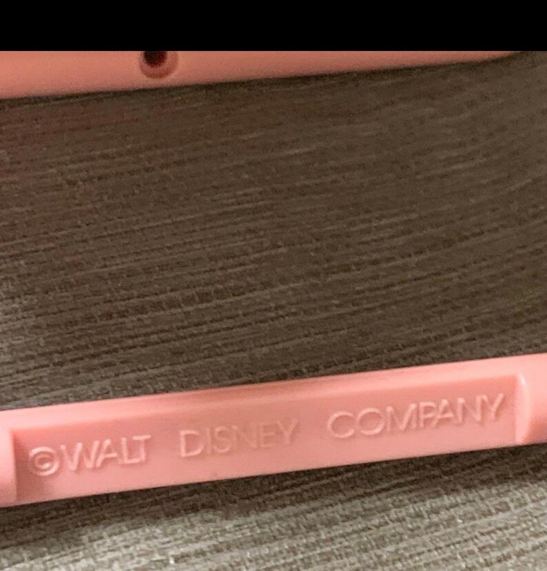 May include: A close-up of a light pink plastic object with the text "WALT DISNEY COMPANY" printed in a darker pink. The object is rectangular and appears to be part of a larger item.