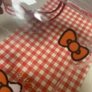 Pode incluir: Saco de pl&aacute;stico transparente com cord&atilde;o vermelho e padr&atilde;o xadrez vermelho e branco. O saco apresenta um design de la&ccedil;o laranja e as palavras "Hello Kitty".