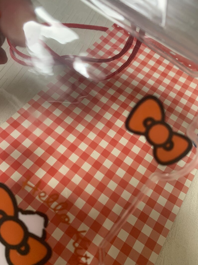 Pode incluir: Saco de pl&aacute;stico transparente com cord&atilde;o vermelho e padr&atilde;o xadrez vermelho e branco. O saco apresenta um design de la&ccedil;o laranja e as palavras "Hello Kitty".