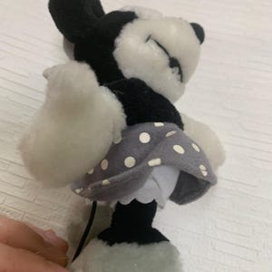 Op de afbeelding: Een Minnie Mouse pop in zwart, wit en grijs. De pop heeft een grijze polkadot rok, witte handschoenen en een zwarte staart. De ogen van de pop zijn gesloten en het lijkt te slapen. De pop is zacht en knuffelbaar.