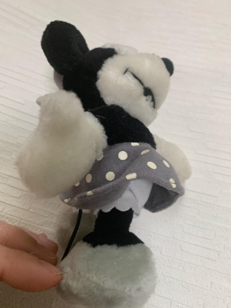 Op de afbeelding: Een Minnie Mouse pop in zwart, wit en grijs. De pop heeft een grijze polkadot rok, witte handschoenen en een zwarte staart. De ogen van de pop zijn gesloten en het lijkt te slapen. De pop is zacht en knuffelbaar.