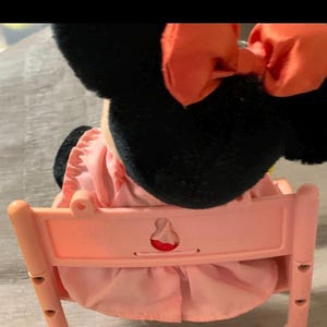May include: A plush Minnie Mouse doll with black ears and a large orange bow. The doll is wearing a pink dress and seated in a light pink plastic chair. The chair has a decorative cutout on the back.