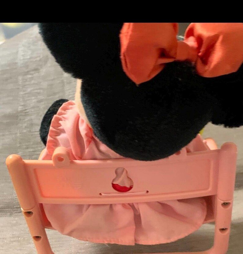 May include: A plush Minnie Mouse doll with black ears and a large orange bow. The doll is wearing a pink dress and seated in a light pink plastic chair. The chair has a decorative cutout on the back.