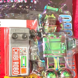May include: A vintage toy robot in its original packaging. The robot is iridescent with a clear helmet and a remote control labeled "CHIBI ROBO". The packaging features Japanese text and a graphic of the robot.
