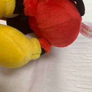 Puede incluir: Un peluche con cuerpo rojo, manos amarillas y detalles negros. El juguete tiene una cabeza negra y una etiqueta de Disney. El juguete est&aacute; hecho de material suave y afelpado.