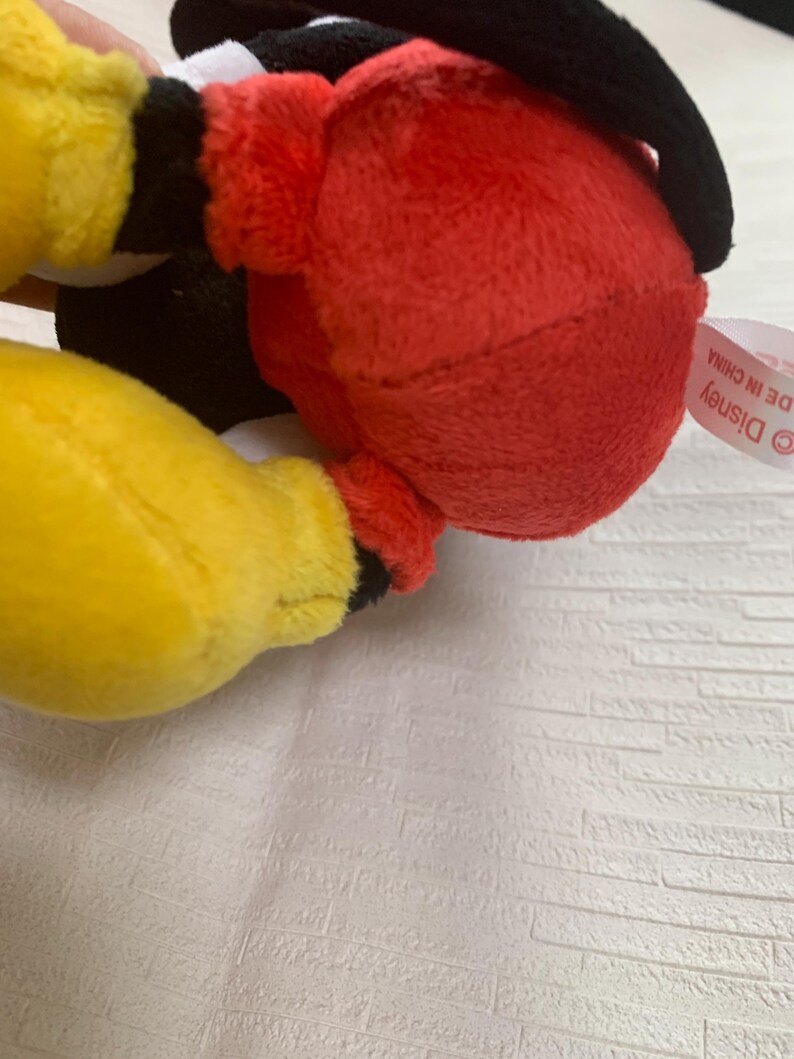 Puede incluir: Un peluche con cuerpo rojo, manos amarillas y detalles negros. El juguete tiene una cabeza negra y una etiqueta de Disney. El juguete est&aacute; hecho de material suave y afelpado.
