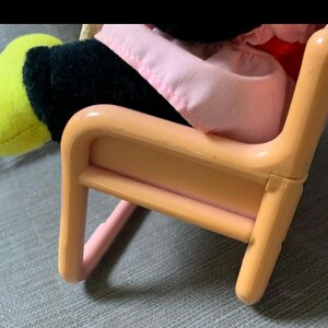 May include: A small, peach-colored plastic chair with rounded edges. A plush toy with a black body, yellow hand, and pink clothing is seated in the chair. The chair's design is simple and child-like.