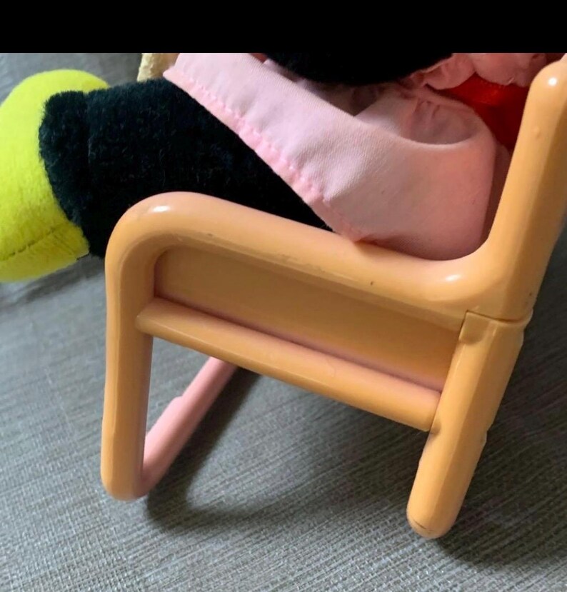 May include: A small, peach-colored plastic chair with rounded edges. A plush toy with a black body, yellow hand, and pink clothing is seated in the chair. The chair's design is simple and child-like.
