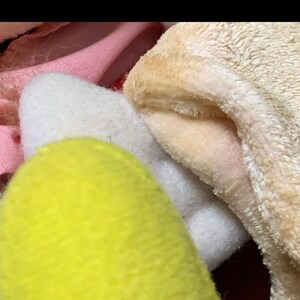 May include: Close-up of soft, plush toys in various colors. A bright yellow, rounded shape is in the foreground, with a white, fluffy shape behind it. A tan, fuzzy fabric is visible on the right, and pink fabric is in the background.