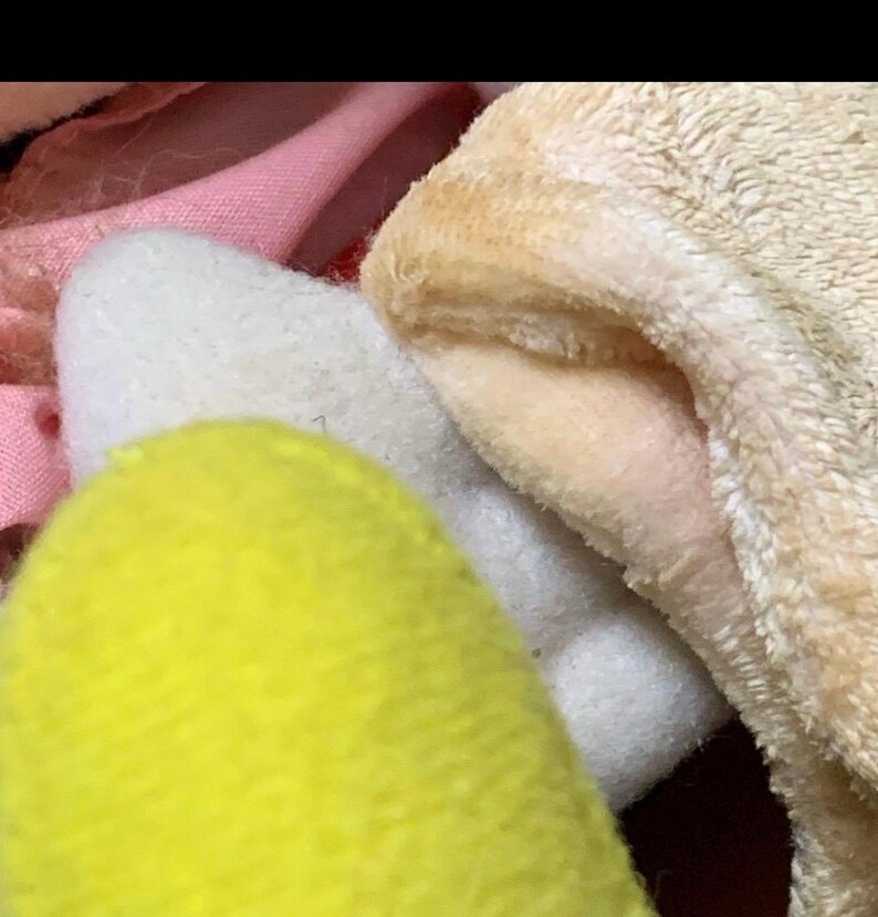 May include: Close-up of soft, plush toys in various colors. A bright yellow, rounded shape is in the foreground, with a white, fluffy shape behind it. A tan, fuzzy fabric is visible on the right, and pink fabric is in the background.