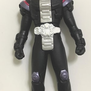 May include: A black action figure of a costumed character with a white and red mask. The figure has a silver chest plate, white belt, and purple accents on the shoulders and knees. The figure is standing upright.