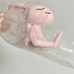 Puede incluir: Una peque&ntilde;a figura de color rosa claro de un conejo con orejas largas y expresi&oacute;n de ojos cerrados, envuelta en una bolsa de pl&aacute;stico transparente y arrugada. El conejo se muestra en posici&oacute;n sentada con sus extremidades visibles a trav&eacute;s.