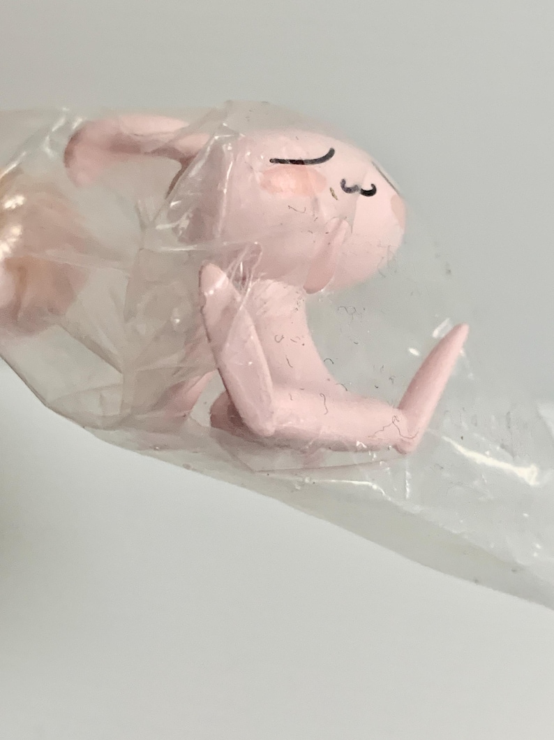 Puede incluir: Una peque&ntilde;a figura de color rosa claro de un conejo con orejas largas y expresi&oacute;n de ojos cerrados, envuelta en una bolsa de pl&aacute;stico transparente y arrugada. El conejo se muestra en posici&oacute;n sentada con sus extremidades visibles a trav&eacute;s.