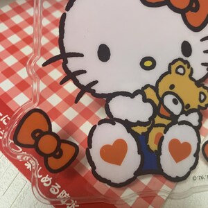Pode incluir: Capa de telefone de pl&aacute;stico transparente com um gato de desenho animado branco com um grande la&ccedil;o laranja e um pequeno ursinho de pel&uacute;cia amarelo. O gato tem olhos pretos e um pequeno nariz amarelo. A capa tem um fundo xadrez vermelho e branco.