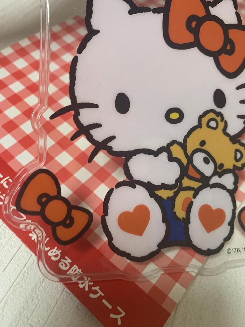 Pode incluir: Capa de telefone de pl&aacute;stico transparente com um gato de desenho animado branco com um grande la&ccedil;o laranja e um pequeno ursinho de pel&uacute;cia amarelo. O gato tem olhos pretos e um pequeno nariz amarelo. A capa tem um fundo xadrez vermelho e branco.