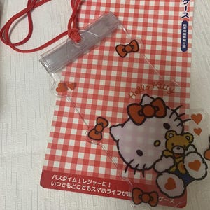Pode incluir: Uma capa de telefone transparente e &agrave; prova d'&aacute;gua com um cord&atilde;o vermelho, com gr&aacute;ficos Hello Kitty e um fundo xadrez vermelho e branco. A capa tem la&ccedil;os vermelhos e detalhes de cora&ccedil;&atilde;o. O texto na embalagem diz "Hello Kitty" e texto japon&ecirc;s.