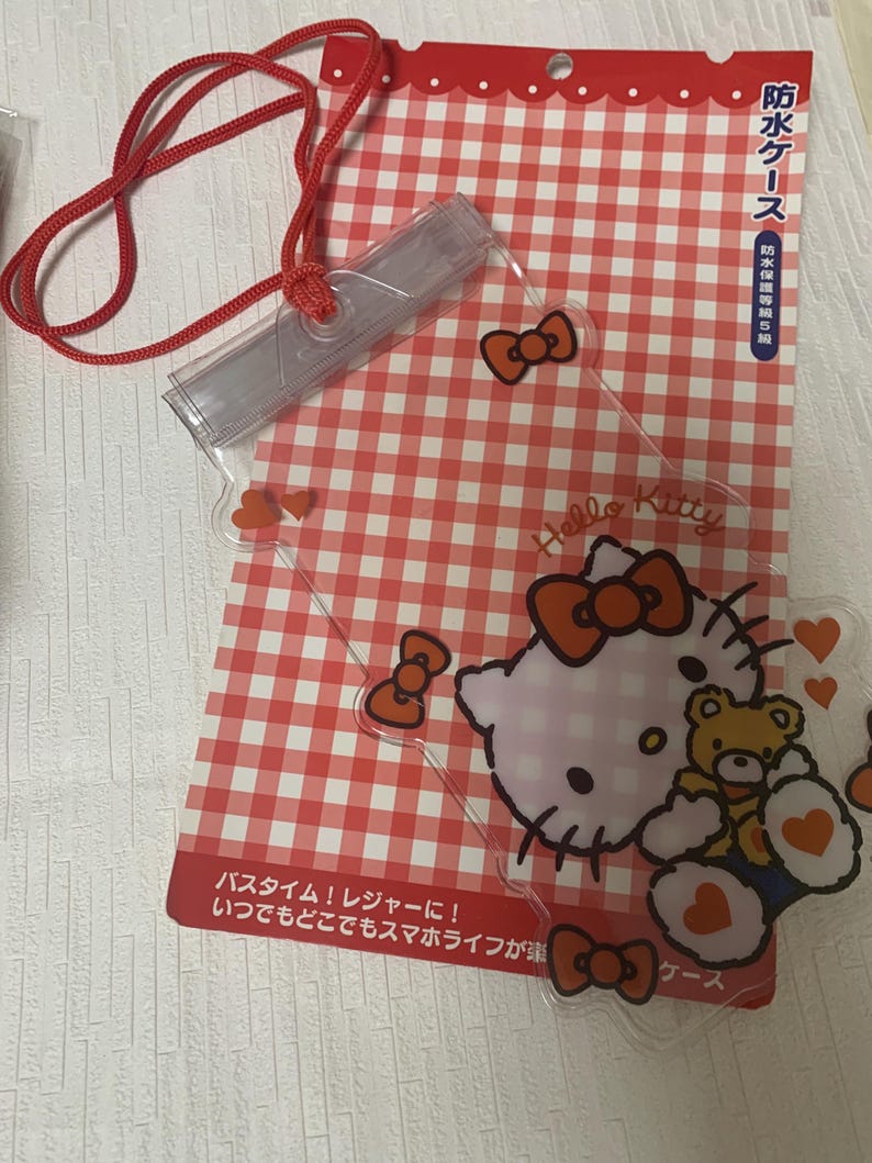 Pode incluir: Uma capa de telefone transparente e &agrave; prova d'&aacute;gua com um cord&atilde;o vermelho, com gr&aacute;ficos Hello Kitty e um fundo xadrez vermelho e branco. A capa tem la&ccedil;os vermelhos e detalhes de cora&ccedil;&atilde;o. O texto na embalagem diz "Hello Kitty" e texto japon&ecirc;s.