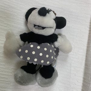 Op de afbeelding: Een Minnie Mouse pluche pop in zwart, wit en grijs. De pop heeft een zwarte top, een grijze rok met witte stippen en grijze schoenen. Het gezicht van de pop is wit met zwarte oren en een zwarte neus. Een klassiek Disney-personage.