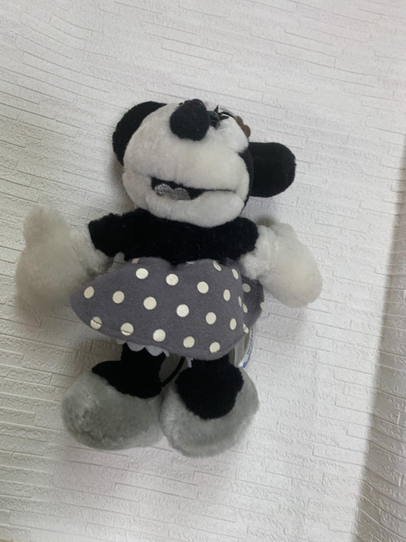 Op de afbeelding: Een Minnie Mouse pluche pop in zwart, wit en grijs. De pop heeft een zwarte top, een grijze rok met witte stippen en grijze schoenen. Het gezicht van de pop is wit met zwarte oren en een zwarte neus. Een klassiek Disney-personage.