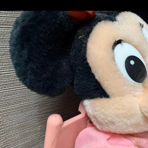 May include: Close-up of a plush Minnie Mouse toy. The toy features black ears, a pink bow, and a light pink face. The eye is white with a black pupil. The toy is in a pink stroller.