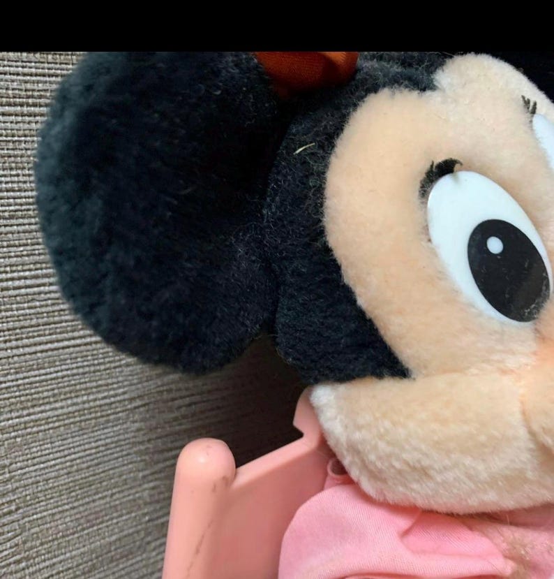 May include: Close-up of a plush Minnie Mouse toy. The toy features black ears, a pink bow, and a light pink face. The eye is white with a black pupil. The toy is in a pink stroller.