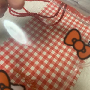 Pode incluir: Uma sacola pl&aacute;stica transparente com um cord&atilde;o vermelho e um padr&atilde;o xadrez vermelho e branco. A sacola apresenta gr&aacute;ficos em forma de la&ccedil;o laranja. O fundo &eacute; uma superf&iacute;cie branca.