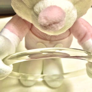 May include: A clear, circular bangle bracelet made of a smooth, transparent material. The bracelet is held by a plush toy with light yellow fur, a pink nose, and white fabric accents on the arms and feet, wearing a light pink garment.