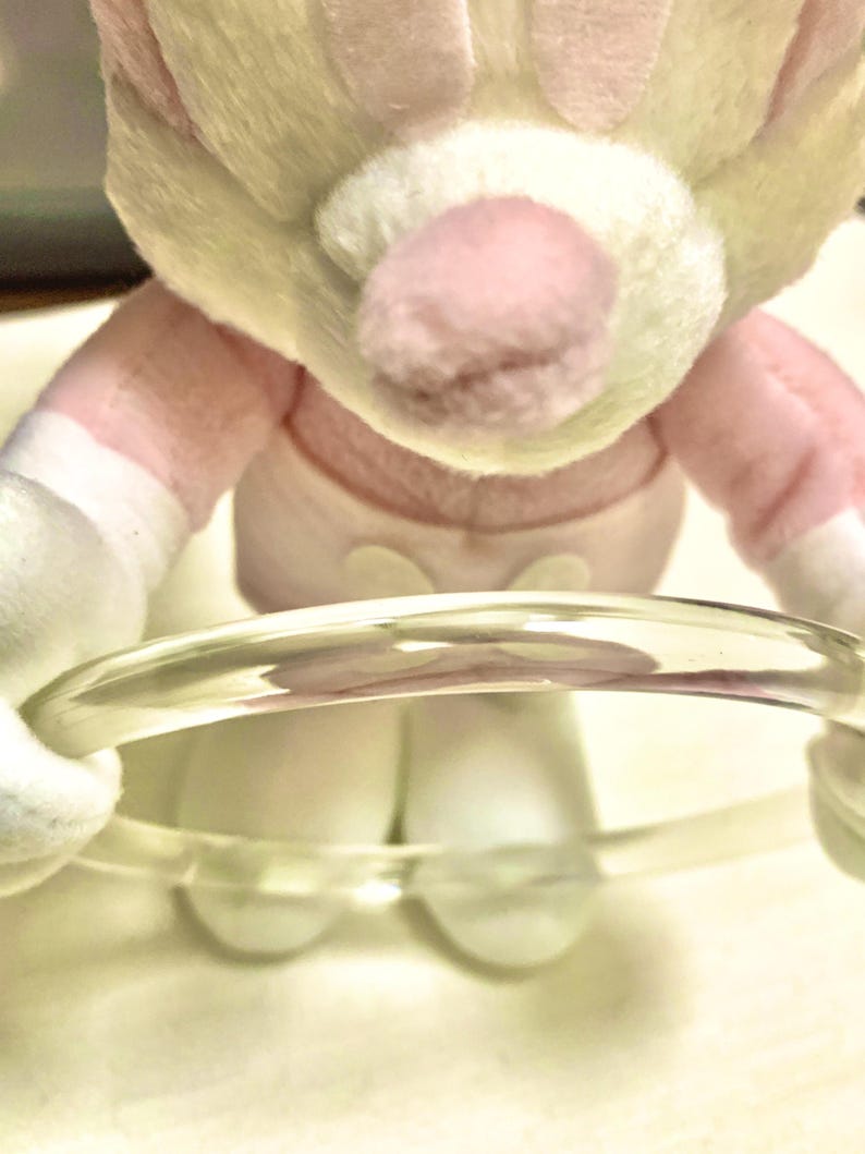 May include: A clear, circular bangle bracelet made of a smooth, transparent material. The bracelet is held by a plush toy with light yellow fur, a pink nose, and white fabric accents on the arms and feet, wearing a light pink garment.