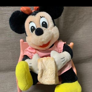 May include: A Minnie Mouse plush toy, featuring a pink dress, black ears, and yellow shoes. The plush is seated on a pink chair and holding a beige cloth. The bow on her head is orange.