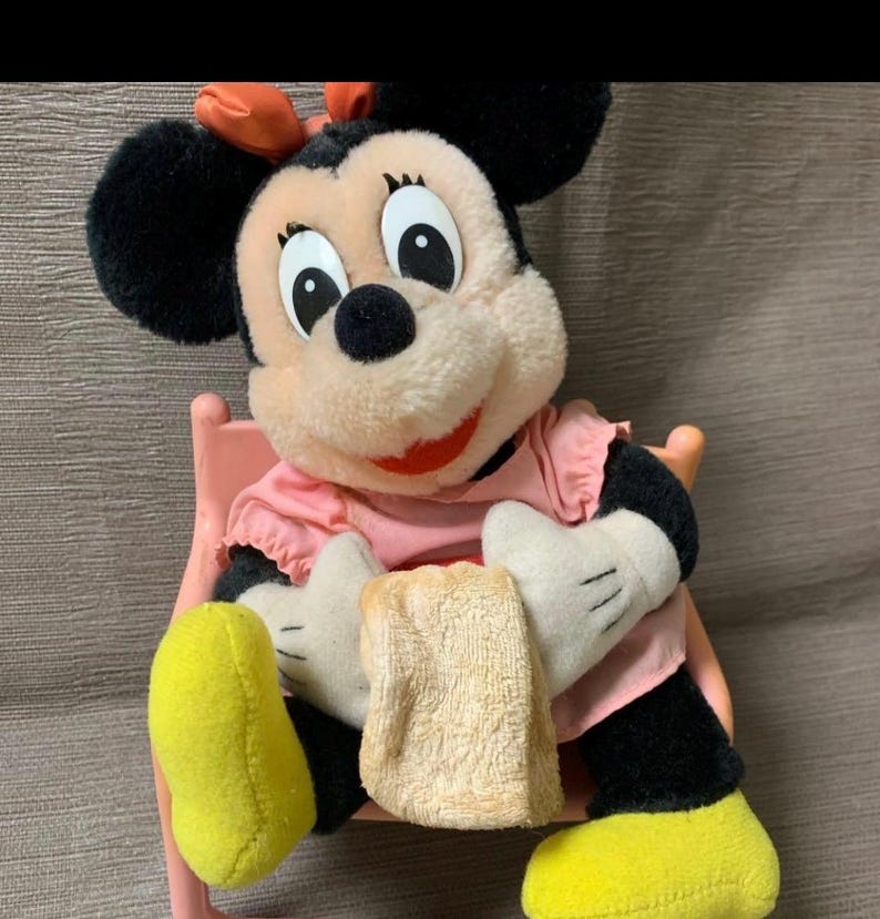 May include: A Minnie Mouse plush toy, featuring a pink dress, black ears, and yellow shoes. The plush is seated on a pink chair and holding a beige cloth. The bow on her head is orange.