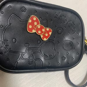 May include: A black rectangular crossbody bag featuring an embossed Hello Kitty outline and small flower and bubble motifs. The bag includes a prominent red and cream polka-dot bow emblem attached to the front and a gold-tone zipper closure.