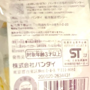 May include: A white product information label with black Japanese text, a barcode, and the ST 22 safety mark. The label includes contact details for Bandai, a 3-year age rating, and a serial number, all printed on a rectangular paper tag.