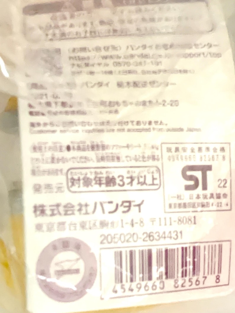 May include: A white product information label with black Japanese text, a barcode, and the ST 22 safety mark. The label includes contact details for Bandai, a 3-year age rating, and a serial number, all printed on a rectangular paper tag.
