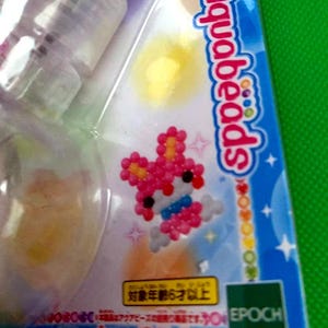 May include: A package of Aquabeads, a craft activity set. The packaging is blue and white, with a pink and white bead design of a rabbit. The text "Aquabeads" is visible. The package also includes the Epoch logo.