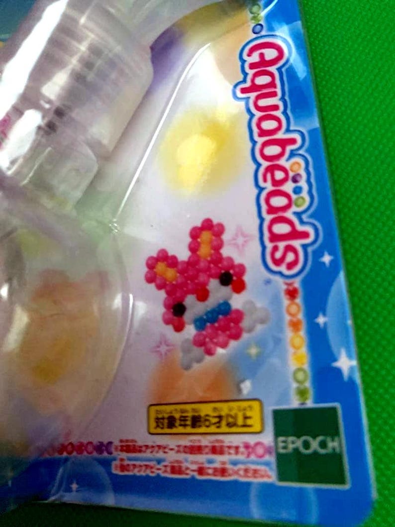 May include: A package of Aquabeads, a craft activity set. The packaging is blue and white, with a pink and white bead design of a rabbit. The text "Aquabeads" is visible. The package also includes the Epoch logo.