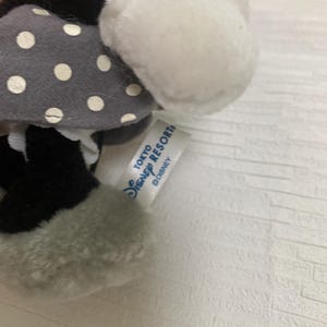 Op de afbeelding: Een pluche Minnie Mouse pop met een grijze jurk met witte stippen, zwarte schoenen en witte handschoenen. De pop heeft een wit gezicht en oren. Een label leest "TOKYO DISNEY RESORT" en het Disney-logo.