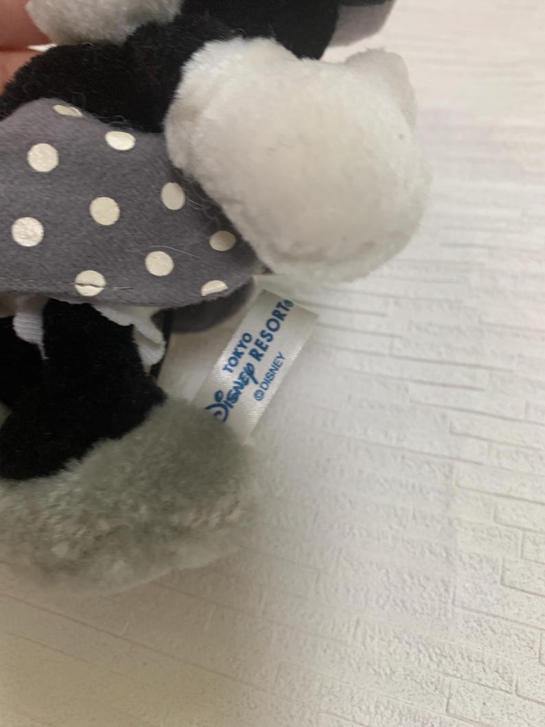 Op de afbeelding: Een pluche Minnie Mouse pop met een grijze jurk met witte stippen, zwarte schoenen en witte handschoenen. De pop heeft een wit gezicht en oren. Een label leest "TOKYO DISNEY RESORT" en het Disney-logo.