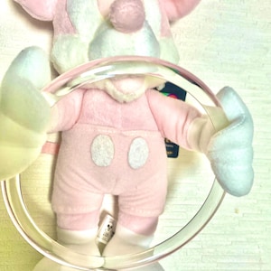 May include: A plush toy character with pastel pink and light blue fur, featuring large rounded ears, a round nose, and two white oval buttons on the torso. The character is holding a large, clear, circular plastic ring in its hands.