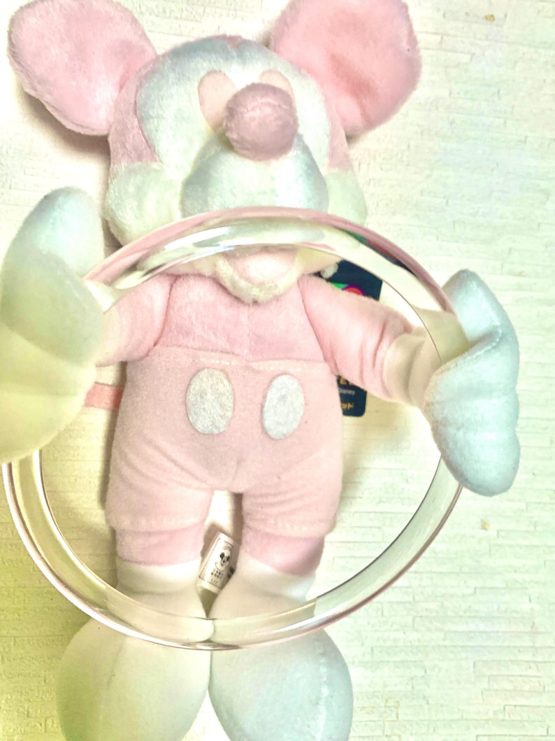 May include: A plush toy character with pastel pink and light blue fur, featuring large rounded ears, a round nose, and two white oval buttons on the torso. The character is holding a large, clear, circular plastic ring in its hands.