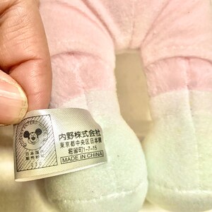 May include: A plush toy with light pink fabric legs and white feet, featuring a white rectangular tag with black text. The tag displays a Mickey Mouse icon, the text '内野株式会社', '東京都中央区日本橋', '堀留町1-7-15', and 'MADE IN CHINA'.