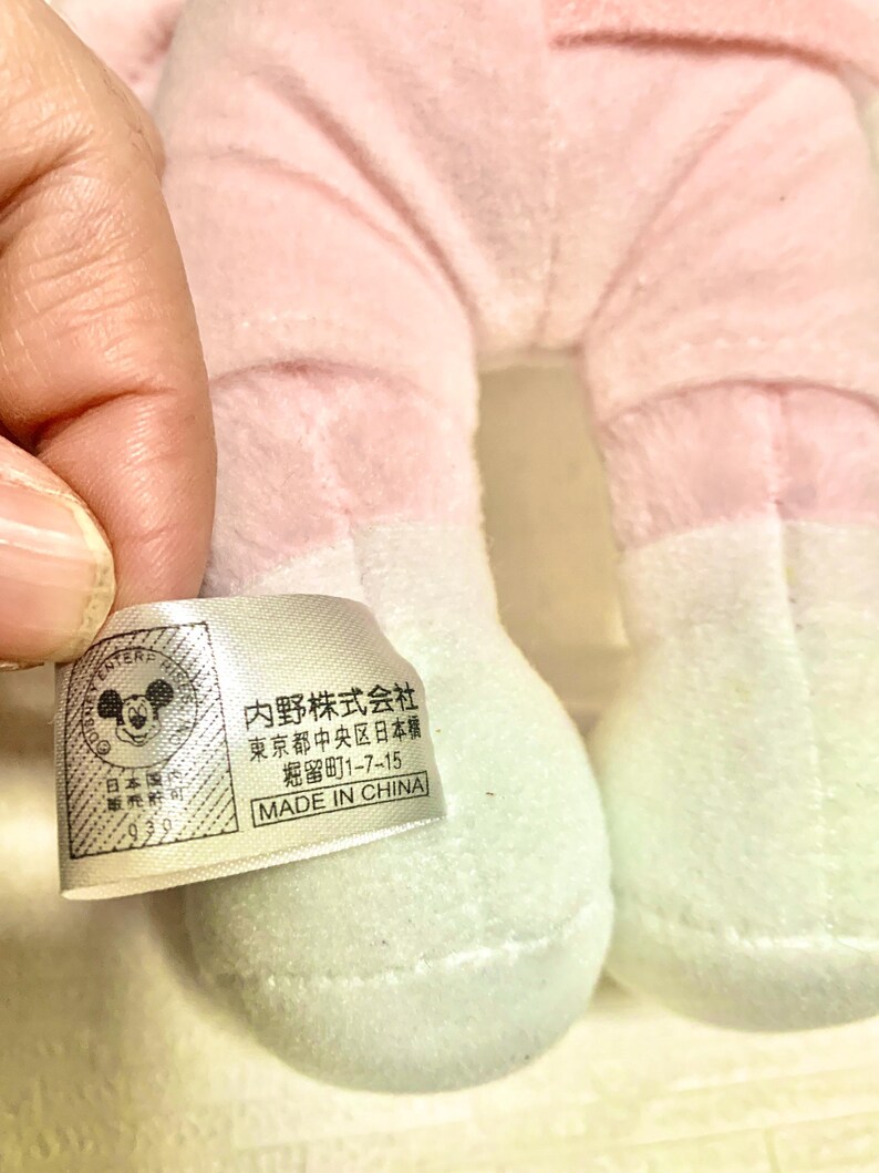 May include: A plush toy with light pink fabric legs and white feet, featuring a white rectangular tag with black text. The tag displays a Mickey Mouse icon, the text '内野株式会社', '東京都中央区日本橋', '堀留町1-7-15', and 'MADE IN CHINA'.