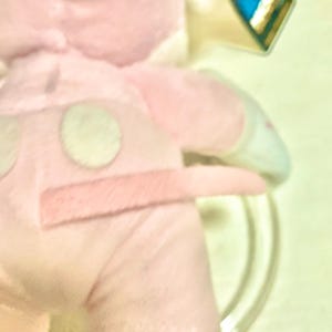 May include: A light pink plush toy featuring a soft fabric texture, a small tail, and two circular patches on the back. The toy includes a small, colorful tag attached to the upper portion, displaying a red shape against a blue background.