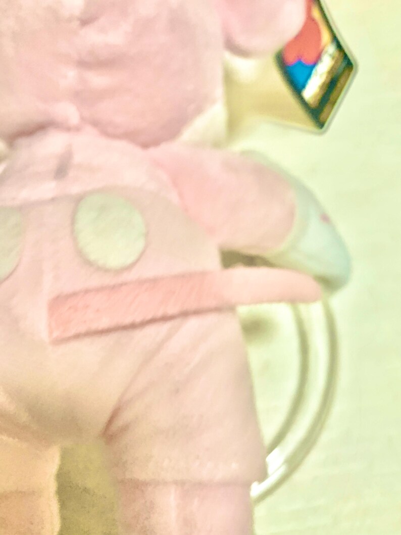 May include: A light pink plush toy featuring a soft fabric texture, a small tail, and two circular patches on the back. The toy includes a small, colorful tag attached to the upper portion, displaying a red shape against a blue background.