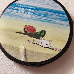Puede incluir: Un estuche redondo con borde negro que presenta una escena de playa impresa con una sandía cortada y una criatura blanca con ojos rojos. El texto Puella Magi Madoka Magica está impreso en blanco en la esquina superior izquierda del diseño.